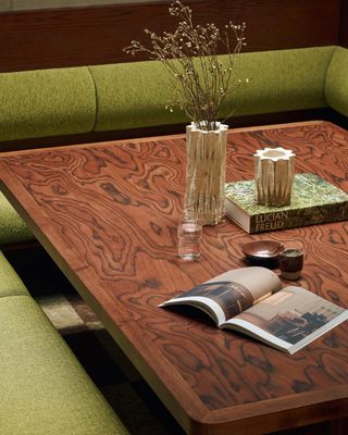 Close-up image of a wooden dining table that has a zebra print wood grain. There is olive green booth seating around the table and a clear glass vase with stems in it